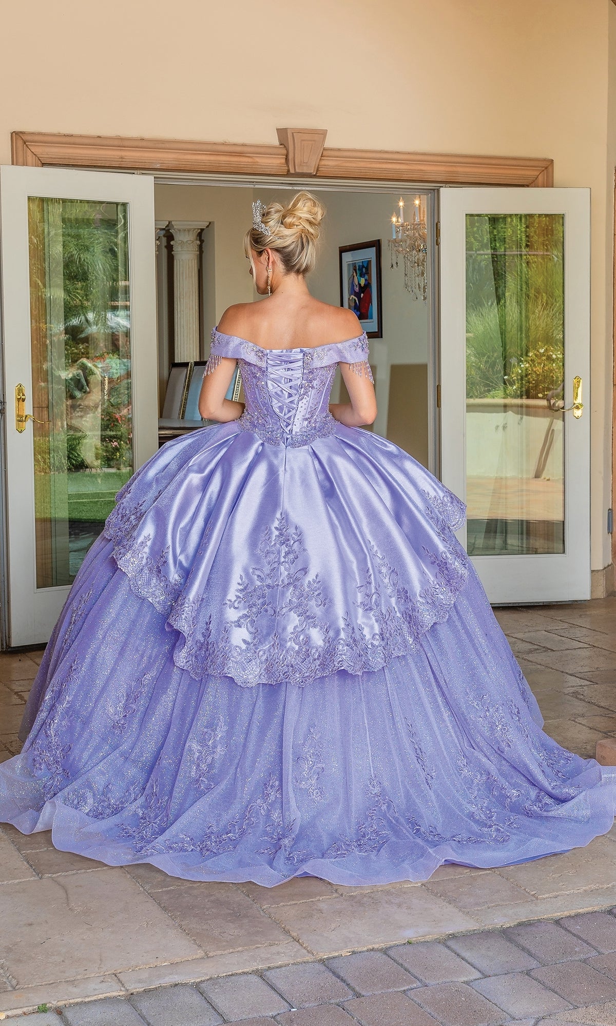 A woman in an off the shoulder Quinceanera Dress 1679 by Dancing Queen with lace details stands outside open double doors, facing away from the camera. The pastel purple gown adds a dreamy touch to the elegant scene.